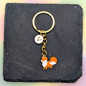 May include: Gold-tone keychain with a fox charm. The fox is orange and white with black accents. The keychain also features a small white charm with a gold letter "V". The keychain is on a black surface.