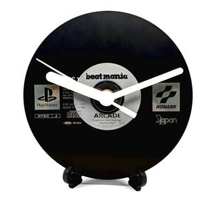 May include: A black CD clock with white hands, featuring the text "beatmania" and PlayStation and Konami logos. The clock is mounted on a black stand. A unique decorative item, the clock measures approximately 12 cm in diameter.
