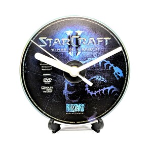 May include: A clock made from a StarCraft II DVD, featuring the game's logo and a blue and white design. The clock has white hands and is mounted on a black stand. The clock's diameter is approximately 12 cm.