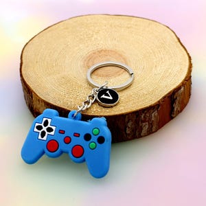 May include: A blue video game controller keyring with red, green, and white details. The keyring features a silver ring and a charm with the letter "V" on a black disc. The controller is placed on a wooden slice.