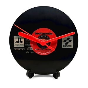 May include: A black vinyl record clock with red hands and a red centre label that reads "beat mania." The clock features the PlayStation and Konami logos. The clock is supported by a black stand.