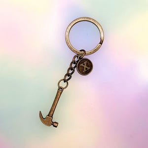 May include: Bronze-toned metal keychain featuring a hammer charm and a circular charm with the letter "X". The keychain has a large ring for keys and a chain connecting the charms. The background is a soft gradient of pastel colors.
