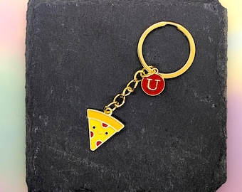 May include: A gold-colored keychain featuring a pizza slice charm with a smiling face and a red letter "U" charm. The pizza slice has yellow crust and toppings. The keychain is on a black surface.