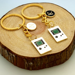 May include: Two gold keychains with a game controller charm and initial charms. The charms are black and white with green accents. The keychains are on a wooden surface.