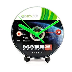 May include: A clock made from an Xbox 360 Mass Effect Disc 1. The clock face features the game's logo and a space scene. It has bright green clock hands and is mounted on a black stand.