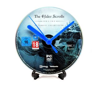 May include: A clock made from a video game disc, with the title "The Elder Scrolls Online: Tamriel Unlimited." The clock face is blue and grey, with blue hands. The clock is supported by a black stand. The disc has a diameter of approximately 12 cm.