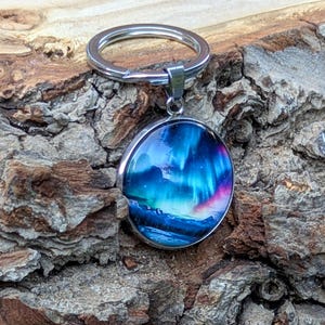 May include: A circular keyring featuring a vibrant image of the aurora borealis with swirling shades of blue, purple, and pink light above a dark landscape. The pendant is encased in a polished silver-toned metal frame and attached to a standard split ring.