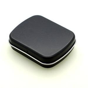 May include: A small, black, rectangular tin with rounded corners and a silver trim. The lid is closed, and the tin sits on a white surface. The tin appears to be empty and ready for use.