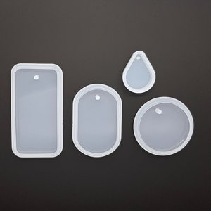 May include: Four clear silicone moulds in different shapes: a rectangle, an oval, a teardrop, and a circle. Each mould has a raised border and a small hole for hanging. The moulds are arranged on a dark grey surface.
