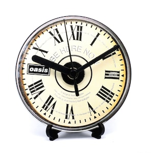 May include: A white and black clock with Roman numerals and the words "Be Here Now" and "Oasis" on the face. The clock is on a black stand.