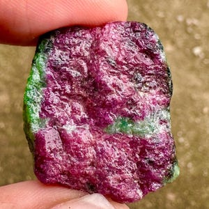 以下が含まれることがあります： 深い赤色の原石ルビー。緑と黒の縞模様がアクセントになっています。自然石は指で挟まれ、その粗い質感の表面が強調されています。このユニークな鉱物標本は、天然ルビーの美しい例です。