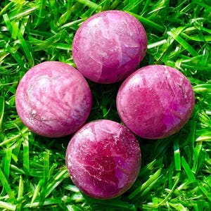 Könnte beinhalten: Vier polierte, runde, rosa Edelsteine mit unterschiedlichen Farbtönen sind auf einem Bett aus grünem Gras angeordnet. Die Edelsteine haben eine glatte, reflektierende Oberfläche, und die Grashalme sind lang und dünn.