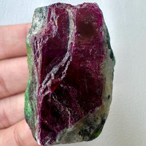May include: A raw ruby gemstone, displaying a rich, deep red colour with patches of green and grey. The natural stone has a textured surface and an irregular shape, highlighting its unpolished state. Ideal for jewellery making or as a collector's item.