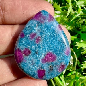 Puede incluir: Una gema en forma de lágrima con una base azul vibrante salpicada de inclusiones magenta profundas. La piedra pulida se sostiene en una mano, con follaje verde en el fondo. La superficie de la gema es lisa y reflectante, mostrando un patrón natural único.