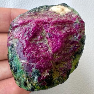 May include: A raw ruby gemstone, displaying a rich magenta colour. The stone has a textured surface with green and black patches, and a small white area. The ruby is held in a hand against a white backdrop.