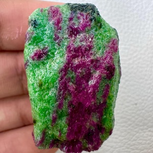 Könnte beinhalten: Ein roh geschnittener Edelstein mit einer lebendigen Farbmischung. Der Stein hat eine Basis aus hellem Grün, mit Streifen und Flecken in tiefem Magenta und einem Hauch von Schwarz. Die Textur erscheint rau und natürlich.