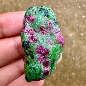 May include: A raw gemstone with a green matrix speckled with magenta and black inclusions. The stone's surface is uneven, displaying its natural composition. The gemstone is held in a hand.