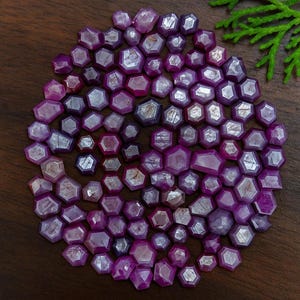 May include: A collection of faceted, deep magenta-colored gemstones, arranged in a circular pattern on a dark wooden surface. The gems have a hexagonal shape and varying sizes. A sprig of green foliage is in the upper right corner.