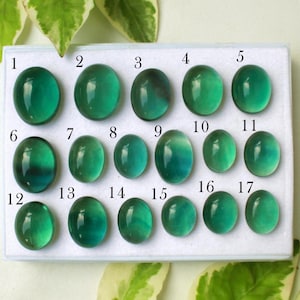 Può includere: Una collezione di diciassette pietre preziose verdi lucide, di forma ovale, disposte in un contenitore rettangolare bianco. Le pietre preziose variano leggermente per colore e dimensione, con una finitura lucida. Numerate da 1 a 17.