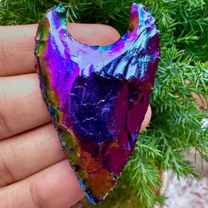 Peut inclure: Une pierre irisée en forme de pointe de flèche avec une surface polie. La pierre affiche un spectre de couleurs, notamment le violet, le bleu, le vert et l'or. Les bords ont un aspect rugueux et texturé. La pierre est tenue sur un fond de feuillage vert.
