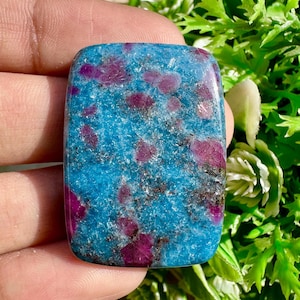 Puede incluir: Una gema rectangular con una base turquesa vibrante, salpicada de inclusiones magenta oscuro y grises. La piedra pulida tiene bordes redondeados y una superficie lisa. La gema se sostiene en una mano, con follaje verde en el fondo.