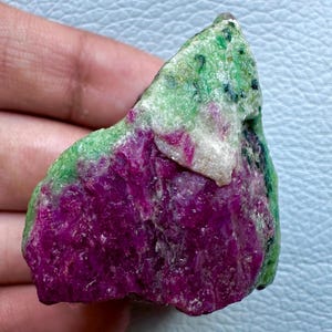 Peut inclure: Une pierre brute de rubis et de zoïsite. La pierre présente une teinte magenta vibrante avec des taches vertes et blanches. Le minéral brut est tenu dans une main, mettant en valeur sa texture naturelle et non polie. Ce spécimen unique est un exemple frappant de beauté minérale naturelle.