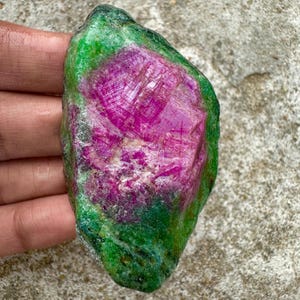Op de afbeelding: Een ruw geslepen edelsteen met een levendig magenta centrum omgeven door een groene buitenlaag. De steen heeft een natuurlijke, ongepolijste uitstraling, die zijn ruwe schoonheid laat zien. De kleuren zijn rijk en verzadigd, wat de unieke kenmerken van het mineraal benadrukt.