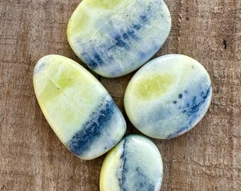 May include: Four polished stones with a mix of colors. The stones have a base color of yellow-green with blue and white marbling. The stones are oval and rounded, and are displayed on a wooden surface.