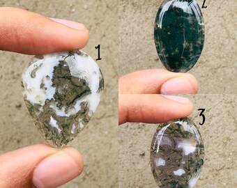 May include: Three polished gemstones are displayed. Gemstone 1 is pear-shaped with white and green patterns. Gemstone 2 is oval-shaped with dark green patterns. Gemstone 3 is oval-shaped with brown and white patterns. Each stone is held between fingers.