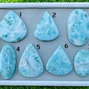 Puede incluir: Una colección de siete piedras preciosas de larimar pulidas en varias formas, incluyendo lágrima, triangular y ovalada. Las piedras muestran una gama de tonos azul claro con patrones blancos y azul oscuro, sobre un fondo gris dentro de un marco blanco.