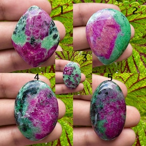 May include: A collection of five ruby zoisite gemstones. The stones display a mix of colors, including deep pink, green, and black. Each stone has a unique shape and pattern, showcasing the natural beauty of the minerals.