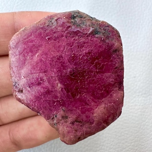 Puede incluir: Una gema de rubí en bruto de color magenta intenso. La superficie tiene un aspecto texturizado con patrones naturales visibles y algunas inclusiones oscuras. Los bordes son de un color rosa polvoriento más claro.