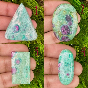 May include: Four polished gemstones in different shapes: a triangle, an oval, a rectangle, and an elongated oval. The stones are mainly teal with bright pink and red inclusions, set against a backdrop of green moss.