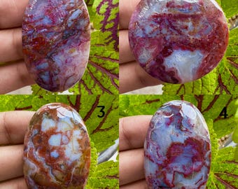 May include: Four polished gemstones with distinctive patterns and colours. The stones display a blend of red, blue, and white shades, with intricate veining and swirls. The shapes vary, including oval and round forms, showcasing natural stone variations.