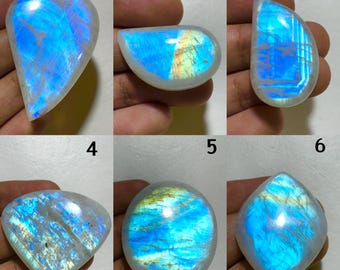 May include: Six polished moonstone gemstones with iridescent blue and white flashes. Each stone has a unique shape, including teardrop, oval, and rounded square. The stones are held in a hand, showcasing their natural beauty and potential for jewelry making.