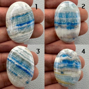 May include: Four oval-shaped gemstones with a polished finish. Each stone features a mix of white, blue, and gray colors, with unique patterns of stripes and swirls. The stones are held in a hand, showcasing their size and detail.