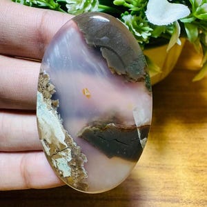 May include: An oval-shaped gemstone with a polished surface. The stone features a blend of colors, including brown, pink, white, and green, with a marbled pattern. The gemstone has a natural, earthy appearance, with a smooth, glossy finish.