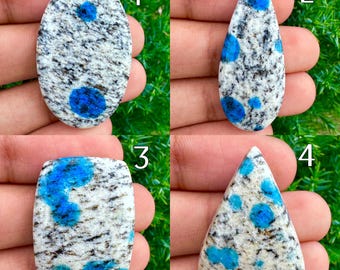 May include: Four K2 stone cabochons in various shapes: oval, teardrop, rectangular, and triangular. Each stone features a white and gray base with vibrant blue spots and inclusions, showcasing natural patterns. These polished stones are ideal for jewelry making.