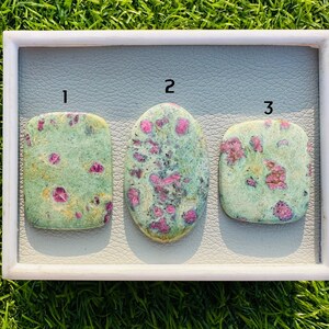 May include: Three polished ruby in zoisite stones in a white tray. The stones are a mix of green and pink, with the pink ruby crystals scattered throughout the green zoisite. The shapes are rectangular, oval, and square.