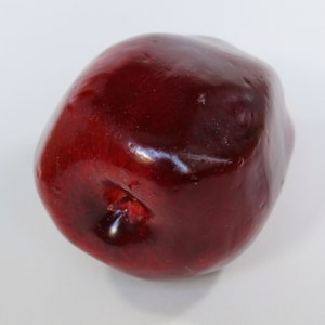 May include: A single, glossy, deep red apple with a smooth surface. The apple has a slight indentation at the bottom and is set against a plain white background. The fruit appears to be freshly picked.
