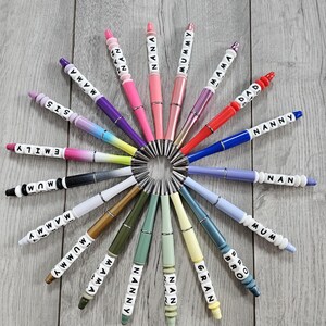 Puede incluir: Una colección de bolígrafos coloridos con diferentes nombres escritos en cuentas blancas. Los bolígrafos están dispuestos en círculo sobre una superficie de madera.