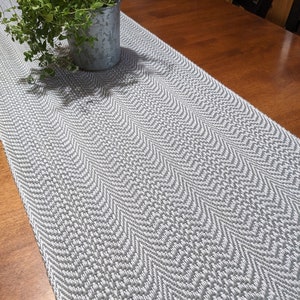 May include: A long, rectangular table runner featuring a woven grey and white chevron pattern. The fabric displays a textured, repeating zigzag design throughout its length, creating a consistent visual rhythm across the surface of the textile.