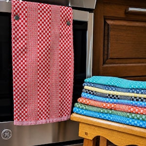 Peut inclure: Un torchon de cuisine à carreaux rouges et blancs est suspendu à la porte d'un four en acier inoxydable. Une pile de torchons de cuisine à carreaux colorés de différentes couleurs est posée sur un tabouret en bois. Les serviettes ont une texture tissée.