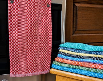 May include: A red and white checkered kitchen towel with two metal snaps hanging from a stainless steel oven handle. A stack of folded kitchen towels in various colors including teal, yellow, black, orange, and green sits on a wooden stool.