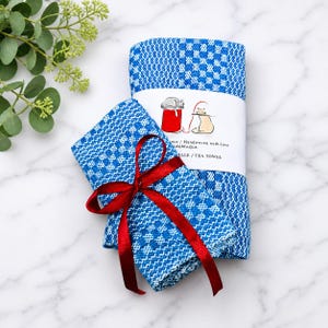 May include: Two blue and white checkered handwoven tea towels. One towel is folded and tied with a red satin ribbon. A white paper band wraps around the towels, featuring a graphic of two cats and the text "Handwoven with love" and "TEA TOWEL".