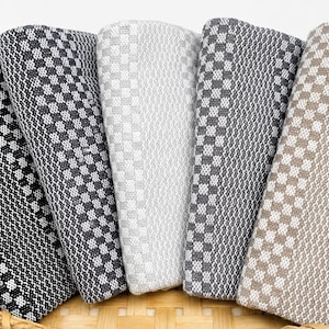 May include: Five folded rectangular textiles with a woven checkered pattern arranged in a fan shape inside a woven wicker basket. The fabrics feature color variations including black, dark gray, light gray, charcoal, and beige with white accents.