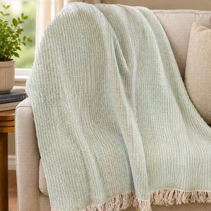 May include: A light green throw blanket with a textured, ribbed design and a cream-colored fringe. The blanket is draped over a beige sofa, creating a cozy and inviting aesthetic. The throw is made of a soft material.