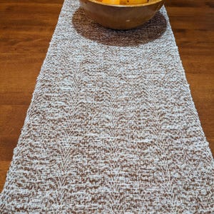 May include: A rectangular table runner featuring a textured, woven pattern in alternating shades of brown and white. A large, round, light-brown wooden bowl filled with several bright orange citrus fruits sits centred on the runner.