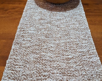 May include: A rectangular table runner featuring a textured, woven pattern in alternating shades of brown and white. A large, round, light-brown wooden bowl filled with several bright orange citrus fruits sits centered on the runner.