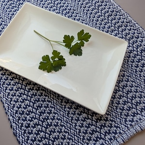 May include: A rectangular white ceramic plate with a smooth, glossy finish sits atop a woven textile featuring a dense, repeating geometric pattern in navy blue and white. A small, fresh sprig of green parsley rests on the centre of the plate.